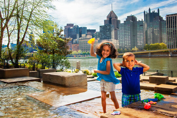 A girl and boy play with toys in the water steps fountain with the city skyline behind them.