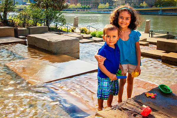 A brother and sister smile and hug while standing in the water steps fountain.