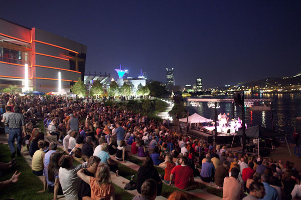 Crowds pack the amphitheater bleachers for an evening concert with the city skyline behind the stage.