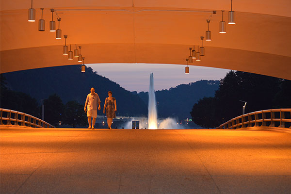 A man and woman walk over the portal bridge toward the camera with the fountain in the background.