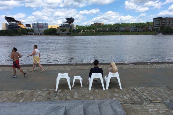 A couple sits in two chairs as a runner and jogger pass by on the riverfront.
