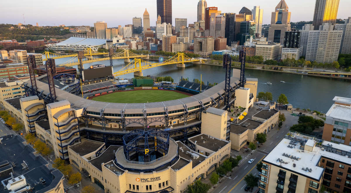 A high-angle view of a modern baseball stadium. The structure is open toward the top and provides a view of suspension bridges crossing a wide river. Across the water is a city skyline composed of both modern and traditional buildings and skyscrapers.