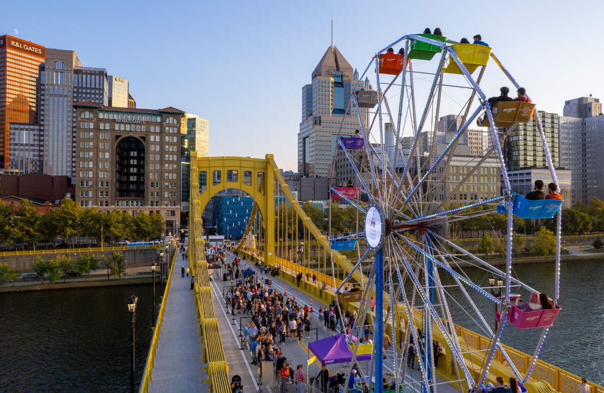 A multicolored Ferris wheel sits in the foreground atop a gold suspension bridge. In the background, large buildings make upi a city skyline.