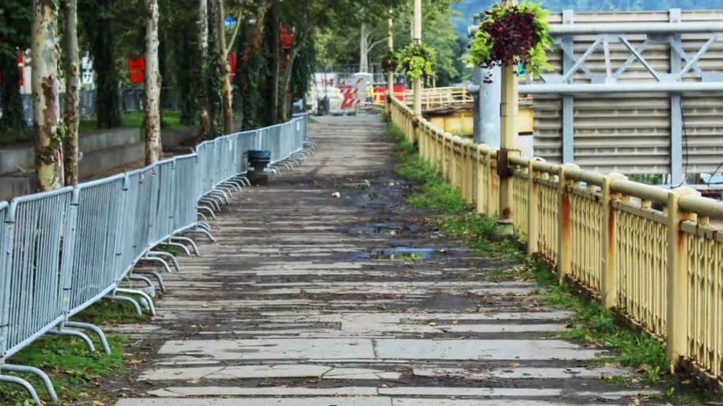 A deteriorated pedestrian path with broken concrete slabs, flanked by temporary metal barriers and a weathered yellow railing, leading to a bridge with industrial features.